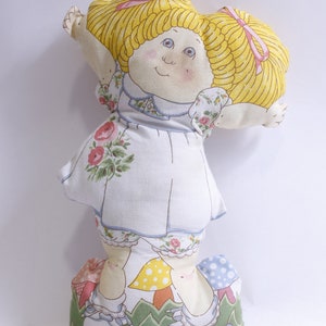 May include: A yellow and white fabric doll with blonde pigtails and a floral dress. The doll is standing on a green and white fabric base with mushrooms.