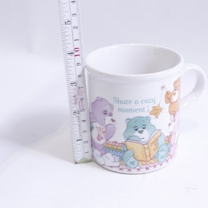 May include: White ceramic mug with a colorful illustration of Care Bears reading a book and the text "Share a cozy moment!"