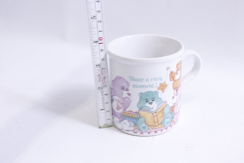 May include: White ceramic mug with a colorful illustration of Care Bears reading a book and the text "Share a cozy moment!"