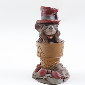 May include: A whimsical figurine of a bear wearing a burgundy top hat, emerging from a brown ice cream cone. The base features red berries and leaves. The bear has a smiling expression.