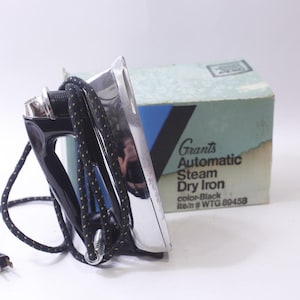 Puede incluir: Plancha de vapor seca automática Grants vintage, negra y plateada, con cable negro. La plancha está junto a su caja original, que tiene el nombre del producto y la información del color impresa.