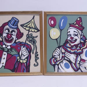 May include: Two framed paintings featuring colorful clown portraits. One clown wears a blue top hat and holds a parasol. The other clown holds balloons. Both have red noses and are smiling. The frames are a light brown color.