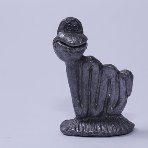 May include: A silver figurine of a cartoon-like creature with a large head and a body made of multiple loops. The creature has a wide smile and large eyes.