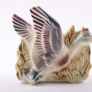 May include: A ceramic planter shaped like a goose in flight with wings spread wide. The goose is painted in shades of purple, green, and brown. The planter is surrounded by green and brown leaves.
