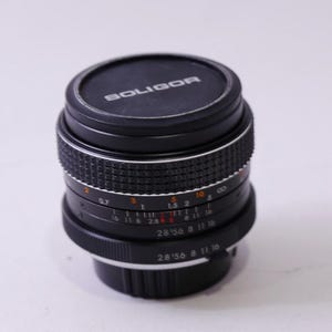 May include: A black Soligor camera lens with a ridged focus ring and aperture settings. The lens has a black cap with the brand name "SOLIGOR" in white lettering. The lens has various numerical markings.