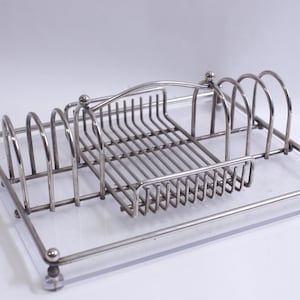Puede incluir: Estante para platos de metal plateado con base de plástico transparente. El estante tiene varios compartimentos para guardar platos y cubiertos.