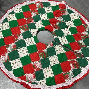 May include: A round Christmas tree skirt with a patchwork design in red, green, and white squares. The skirt has a white lace trim and a center opening for the tree trunk. The squares feature holiday-themed patterns.