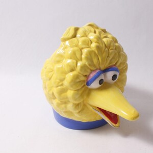 May include: A ceramic Big Bird head figurine featuring bright yellow textured feathers, large white eyes with black pupils, blue eyelids, and a long yellow beak with a red interior. The base of the figurine is a solid blue circular band.