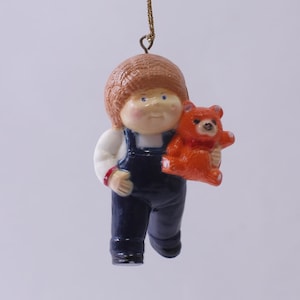 Può includere: Un ornamento natalizio in porcellana che raffigura un bambino con una salopette blu che tiene un orsacchiotto. Il bambino ha i capelli castani e gli occhi azzurri.