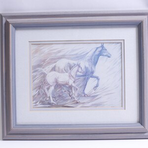 May include: A framed watercolor painting of two white horses, one adult and one foal, running through a field of tall grass. The painting is done in shades of blue and gray, with a soft, ethereal feel.