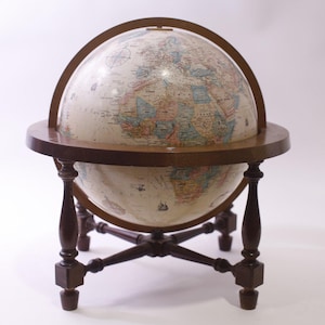 Może przedstawiać: Zabytkowy globus z szczegółową mapą, zamontowany na ciemnej drewnianej podstawie. Globus jest utrzymywany przez drewnianą ramę i spoczywa na czteronożnej podstawie. Kolory globusa obejmują beżowy, niebieski i brązowy.