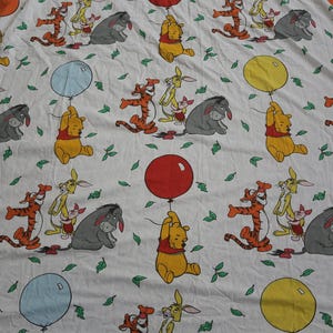 May include: White fabric with a repeating pattern of Winnie the Pooh characters. The design includes Pooh Bear, Tigger, Eeyore, and Piglet, each holding a colourful balloon. Green leaf accents are scattered throughout the design.