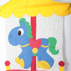 May include: A colorful carousel-themed wall hanging. The design features a blue horse with white polka dots, a red heart, and a green mane and tail. The carousel has a yellow canopy and a red base with yellow accents.