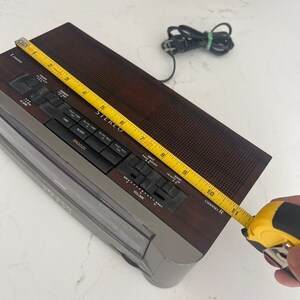 Pu&ograve; includere: Un radio sveglia AM/FM marrone e argento con un display digitale e un lettore di cassette. La radio ha una manopola del volume, un selettore di banda e un timer di spegnimento. La radio misura 25,4 cm di larghezza.