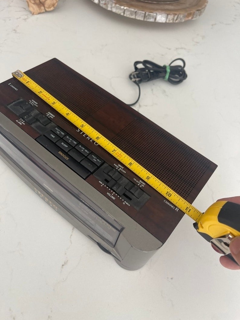 Pu&ograve; includere: Un radio sveglia AM/FM marrone e argento con un display digitale e un lettore di cassette. La radio ha una manopola del volume, un selettore di banda e un timer di spegnimento. La radio misura 25,4 cm di larghezza.