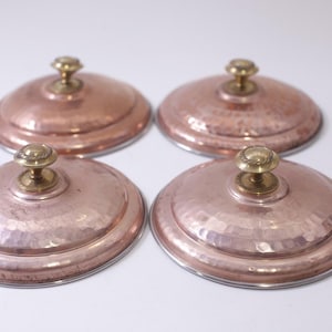 May include: Four copper pot lids with brass knobs. The lids have a hammered texture and a silver rim. The copper has a warm, reddish-brown hue. The knobs are small and round.