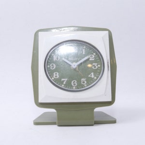 May include: A green and white vintage alarm clock with a square face and a stand. The clock face has black numbers and a green background. The clock says "Security" on the face.