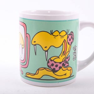 May include: White ceramic mug featuring a cartoon snake in yellow with a pink polka-dot bow. The mug has a light green background with a pink border and a white handle. The mug has the text "S.O.S."