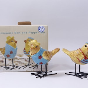 Pode incluir: Conjunto de saleiro e pimenteiro de cerâmica com dois pássaros amarelos de cerâmica vestindo roupas azuis e vermelhas. Os pássaros têm pernas de arame preto e estão em pé sobre uma superfície branca. A caixa diz "Stoneware Salt and Pepper Set".