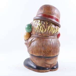Op de afbeelding: Vintage keramieken clownfiguur, met een bruine hoed en rode band, bruine jas en zwarte schoenen. De clown houdt kleurrijke ballen vast. Het beeldje heeft een textuurontwerp en staat tegen een witte achtergrond. De basis heeft de tekst "876 USA".