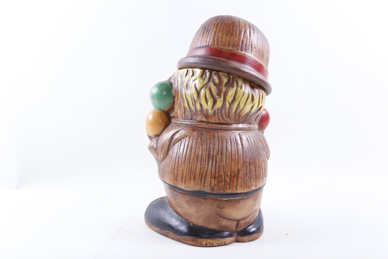 Op de afbeelding: Vintage keramieken clownfiguur, met een bruine hoed en rode band, bruine jas en zwarte schoenen. De clown houdt kleurrijke ballen vast. Het beeldje heeft een textuurontwerp en staat tegen een witte achtergrond. De basis heeft de tekst "876 USA".