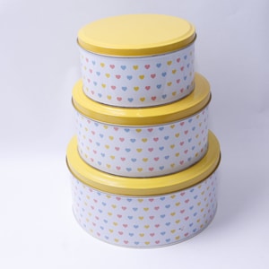 Puede incluir: Una pila de tres latas redondas de metal con tapas amarillas. Las latas son blancas con un patrón de pequeños corazones en rosa, azul y amarillo. Las latas son de diferentes tamaños, la más grande en la parte inferior.