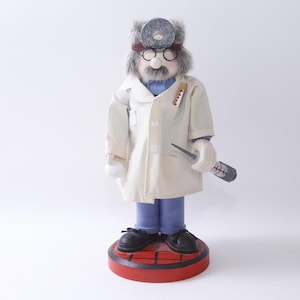 May include: A decorative wooden doctor figurine wearing a white lab coat, glasses, and a stethoscope. The figurine stands on a red and black circular base. The doctor holds a thermometer and a medical instrument.