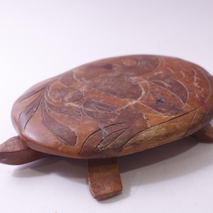 May include: A wooden turtle-shaped decorative item. The brown item has carved details on the top and four legs. The top of the turtle has a carved design of a fish and leaves.