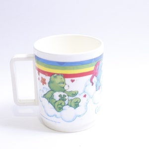 May include: White plastic mug with a rainbow and cloud design featuring a green Care Bear with a four-leaf clover. The Care Bear is sitting on a cloud with a blue Care Bear in the background.