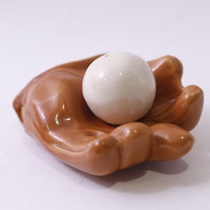 Puede incluir: Un plato de cerámica en forma de mano, con un acabado marrón brillante, sostiene una esfera blanca. La escultura se encuentra sobre un fondo blanco, resaltando el contraste entre el marrón cálido y el blanco frío de la esfera.