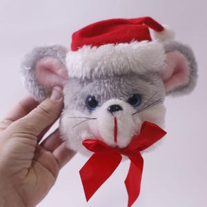 May include: A plush gray mouse head with pink ears, blue eyes, and black whiskers. It wears a red Santa hat with white trim and a red bow. A festive Christmas decoration.