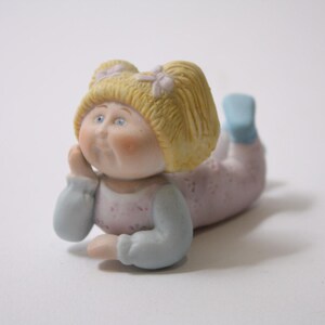 May include: A ceramic figurine of a doll lying down, with blonde hair and blue eyes. The doll is wearing a light pink outfit with blue sleeves and socks. The doll is resting its head on its hand.