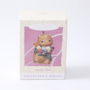 Può includere: Una statuetta di scoiattolo marrone e bianco che tiene un mazzo di fiori colorati. La statuetta fa parte di una collezione Garden Club.