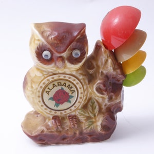 Könnte beinhalten: Eine braune Keramik-Eulenfigur mit großen, weißen Augen. Die Eule hält in ihrem rechten Flügel einen Satz bunter Messlöffel. Die Eule hat ein rundes, weißes Etikett auf ihrer Brust, auf dem "ALABAMA" mit einer roten Rose in der Mitte steht.