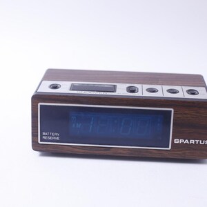 Peut inclure: Un réveil numérique vintage en bois brun avec un affichage numérique bleu affichant l'heure 12:00. L'horloge a un indicateur de réserve de batterie et le nom de la marque "SPARTUS" est imprimé sur le devant.