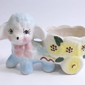 Könnte beinhalten: Ein Keramik-Pflanztopf in Form eines Lammes, das einen Wagen mit gelben Blumen und grünen Blättern zieht. Das Lamm ist hellblau mit rosa Wangen und einer rosa Schleife. Der Wagen ist weiß mit gelben Blumen und grünen Blättern.