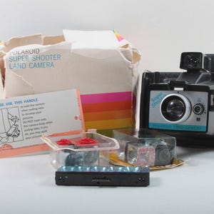 May include: A vintage Polaroid Super Shooter Land Camera with its original box and accessories. The camera is black and silver, with the words "Super Shooter" and "Polaroid Land Camera" printed on the front. The box is beige with colorful stripes. Also included are flash cubes and instructions.