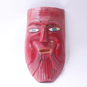 May include: A hand-carved wooden mask painted in a glossy red finish. The mask features a stylized face with a long beard, prominent forehead wrinkles, and almond-shaped eyes with light blue irises and black pupils, all set against a plain background.