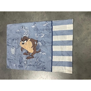 May include: A rectangular pillowcase featuring a blue and white striped panel on the right side. The main section displays a large, colored illustration of the Tasmanian Devil character surrounded by smaller, blue-toned line drawings of Looney Tunes characters.