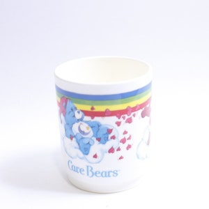 May include: White ceramic mug with a rainbow and hearts design featuring the Care Bears logo and a blue Care Bear with a star on its belly.