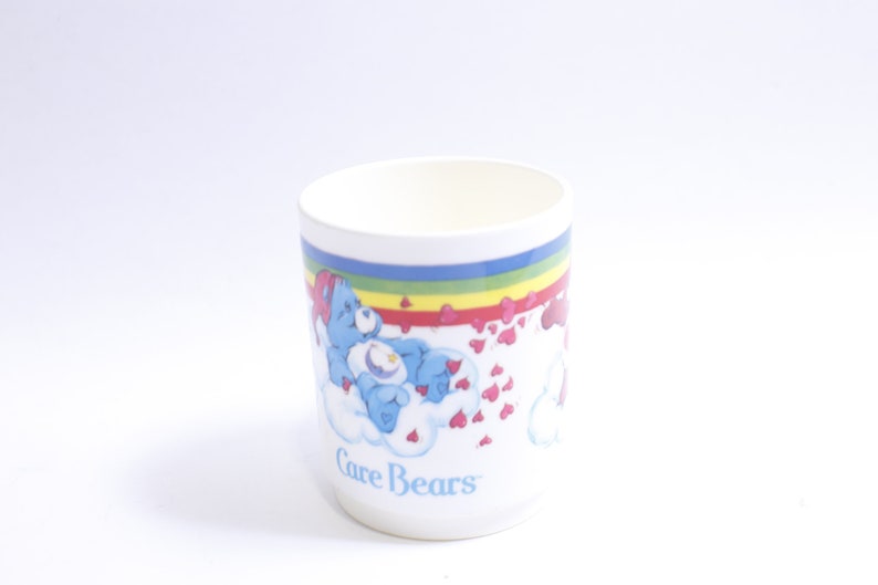 May include: White ceramic mug with a rainbow and hearts design featuring the Care Bears logo and a blue Care Bear with a star on its belly.