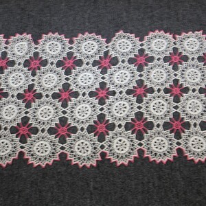 May include: White and pink lace doily with a floral pattern. The doily features a repeating design of white circular shapes with pink floral accents. The edges are scalloped and trimmed in pink. The doily is rectangular in shape.