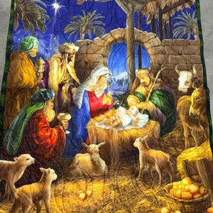Könnte beinhalten: Ein gesteppter Wandteppich, der die Weihnachtsgeschichte darstellt. Das Kunstwerk zeigt das Jesuskind in einer Krippe, umgeben von Maria, Josef, den Heiligen Drei Königen und Tieren. Der Hintergrund ist eine sternenklare Nacht mit Palmen, und die Steppdecke hat einen grünen Rand.
