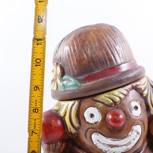 Op de afbeelding: Een vintage keramische clownkoekjespot. De pot is bruin met een rode neus, witte glimlach en geel haar. De clown draagt een bruine hoed met een rode band en houdt kleurrijke ballen vast. De pot is ongeveer 28 cm hoog.