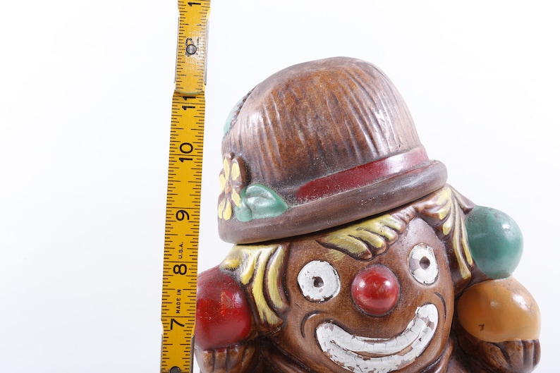 Op de afbeelding: Een vintage keramische clownkoekjespot. De pot is bruin met een rode neus, witte glimlach en geel haar. De clown draagt een bruine hoed met een rode band en houdt kleurrijke ballen vast. De pot is ongeveer 28 cm hoog.