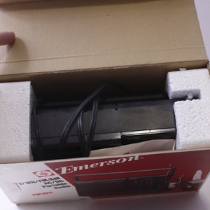 May include: An Emerson TV/WB/FM/AM AC/DC portable radio, model PR3011, is shown inside its original cardboard box. The radio is black and is cushioned with white foam. The box has red and white printed text.