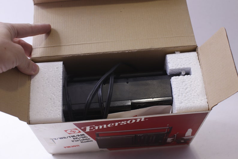 May include: An Emerson TV/WB/FM/AM AC/DC portable radio, model PR3011, is shown inside its original cardboard box. The radio is black and is cushioned with white foam. The box has red and white printed text.
