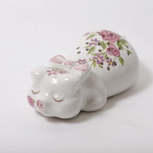 May include: A white ceramic pig figurine in a sleeping pose, adorned with a pink bow and floral accents. The pig's body is decorated with pink roses and purple flowers. The figurine has closed eyes and a smiling expression.