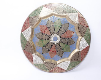 May include: A round brass tray with a decorative design featuring a starburst pattern in shades of blue, green, red, and black. The tray has a raised edge and a smooth surface.