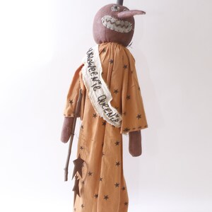 May include: A brown fabric pumpkin doll wearing a tan dress with brown stars and a white sash that says "Halloween Queen". The doll is holding a stick with three brown metal stars attached.